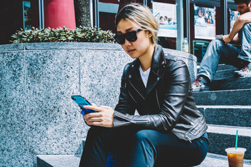 Serious chinese woman in sunglasses checking mail reading messages on mobile sitting on stairs during free time,asian hipster girl in cool apparel spending coffee break outdoors chatting with friend