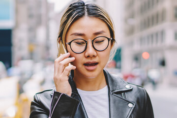 Young asian woman in eyewear walking with ice coffee cup calling taxi contact service operator on...