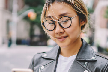Young asian girl in eyewear texting feedback to friend chatting online while spending time outdoors...