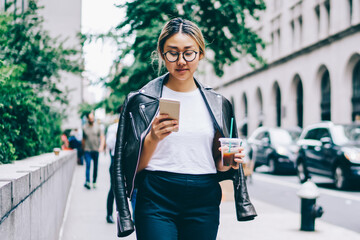 Pensive asian woman walking with coffee to go reading income message on smartphone on street,...