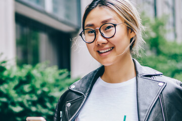 Young asian female tourist in cool apparel and eyewear walking on street using application on phone...