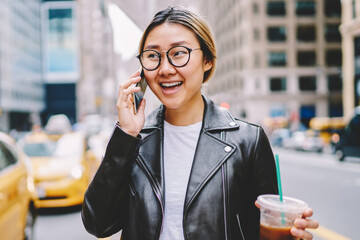 Cheerful female traveler in eyewear talking on mobile in roaming walking on avenue of big city with...