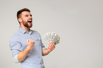 Unexpected win. Young millennial guy showing yes sign with fan of dollars in hand, grey studio...