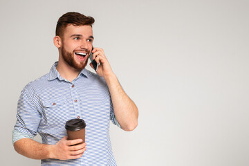 Handsome millennial guy talking on cellphone and holding coffee to go on studio background, copy...