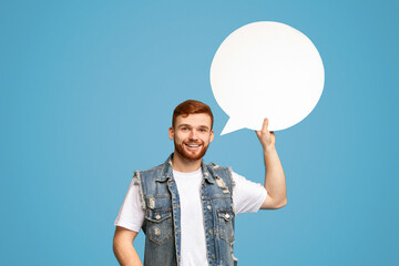 Young bearded guy holding blank speech bubble in one hand and smiling, isolated on blue studio...
