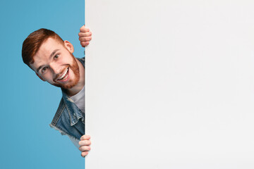 Cheerful young smiling guy looking out of white blank placard ready for text, information or...