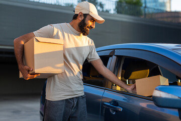 Indian delivery man wearing a beige cap and t-shirt is loading cardboard packages into a car trunk...