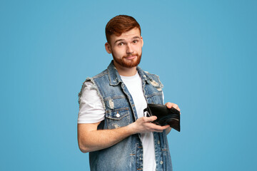 No money for living. Sad millennial man showing his empty wallet to camera on blue background, copy...