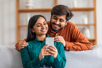 Indian man and woman are sitting together on a couch, both focused on a cell phone in the mans...