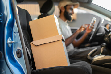 Indian delivery driver is sitting in his parked car, checking his smartphone. Two cardboard boxes...