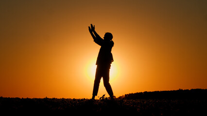 The silhouette of a young fiery woman dances in a field at sunset, highlighted by the orange sky.