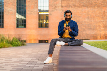 A young Indian man sits outdoors near a modern building, listening to music on his headphones while...