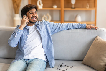 Indian man is sitting on a couch while engaged in a conversation on a cell phone. He appears...