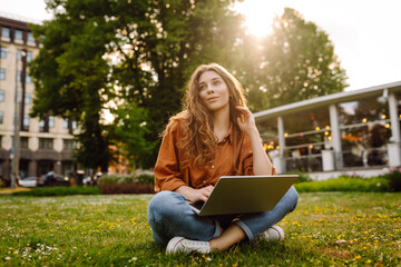 Young woman with laptop sits on the grass in the park on a sunny day.  Online training, remote...