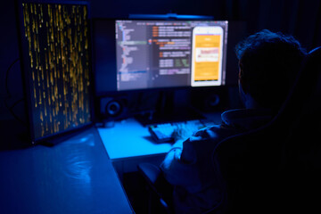 A coder is working late at night on a computer screen, coding and using a smartphone app