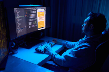 A coder is working late at night on a computer screen, coding and using a smartphone app