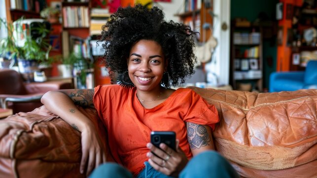 Cheerful woman with curly hair using smartphone while relaxing on comfortable sofa at home