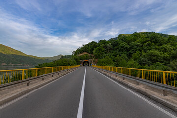 A serene road stretches towards a tunnel entrance, flanked by vibrant greenery and a tranquil lake,...