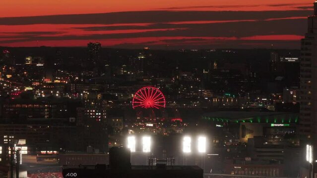 Aerial view of the St. Louis city skyline at sunset, featuring a litup ferris wheel and a baseball stadium with vibrant lights during a game