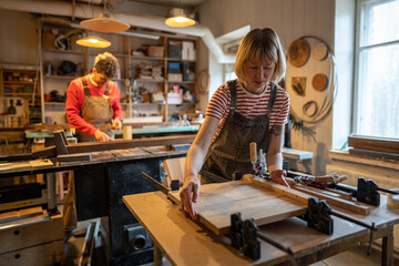 Concentrated female carpenter working on gluing wooden planks together, using clamping brackets and...