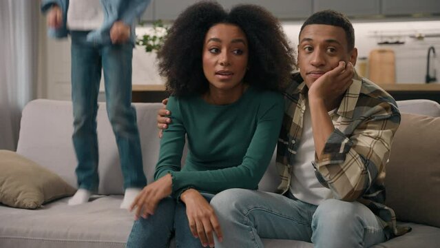 Sad African American family desperate parents hug unhappy mother woman female dissatisfied dad man male sit couch ignore misbehaves little child loud shouting boy active kid son jumping sofa room home