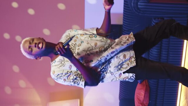 Trandy glamorous cheerful guy in singing into microphone at home party. Creative african american young man having fun, chilling out and dancing at disco neon light illuminated background.