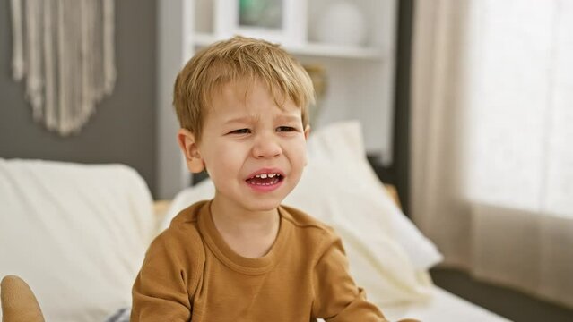 Upset toddler boy crying in a bedroom with white bedding, expressing childhood emotion indoors.
