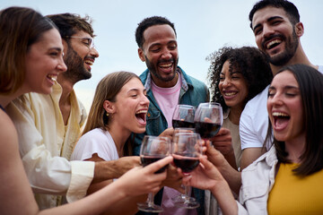 Portrait of multi-ethnic laughing young people toasting glasses red wine outdoor. Gathering of millennial friends together celebrating a party enjoying leisure time on summer holidays