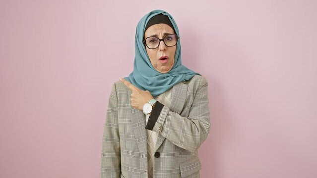 Shocked middle age hispanic woman in hijab, nervously pointing aside with a flustered expression, isolated over pink background.