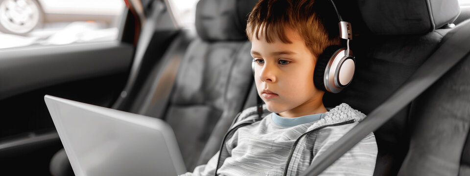 A young boy, headphones on and laptop open, sits transfixed in the backseat of a car, navigating the road ahead as a tech-savvy child,