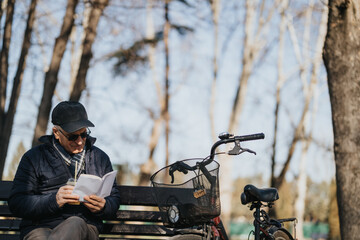 A content elder man with sunglasses and a cap reads a book on a bench, his trusty bike waiting nearby, enjoying a leisurely day outdoors.