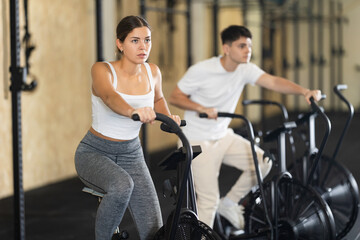 Fototapeta premium Positive girl riding exercise bike for cardio workout during cycling class at modern crossfit gym