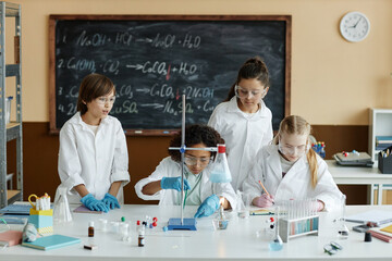 Group of middle school students wearing lab coats and goggles sitting and standing at table in...