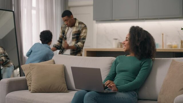Happy African American vacations ethnic family freelance businesswoman workplace smiling woman mother female working typing laptop notebook little child boy son kid dad male man funny dance play home