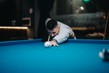 A young man concentrates intensely while aiming a shot in a competitive game of pool in a stylish, modern pool hall.