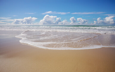 Tide coming in as waves break and roll onto seashore making foamy white patterns across the wet sandy beach, and  horizon with blue cloudy winter sky in background.