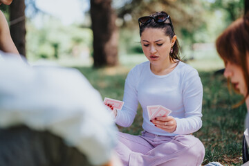 A group of young adults intently playing cards outdoors in a park, capturing a moment of focused...