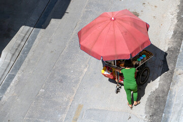 A woman pushes a snack kiosk covered by a large parasol in the sunny street of Ho Chi Minh city. © milkovasa