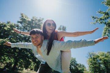 A young man giving a piggyback ride to a woman in a park, both smiling and extending arms like...