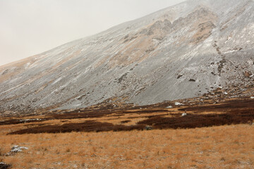 A steep slope of a high mountain with fine gravel on the slopes sprinkled with first snow on the edge of a clearing with yellowed grass on a cloudy autumn day.