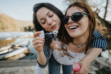 Two cheerful young women enjoying a sunny day outdoors while blowing bubbles by a tranquil lake,...