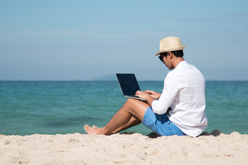 Man traveler use laptop on summer beach blue sky. Asian man casual blue short pants white shirt relax sitting summer beach. Man typing laptop computer holiday business trip freelance work on island