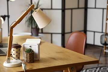 Desk lamp, houseplant and takeaway cup on wooden table in office, closeup