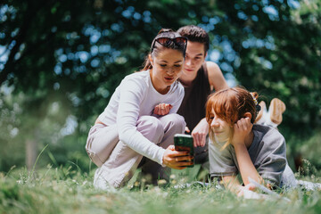 A joyful gathering of young adults in a lush park, centered around sharing content on a smart phone under the shade of trees.