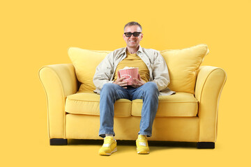 Mature man in 3D glasses with popcorn on sofa watching movie on yellow background