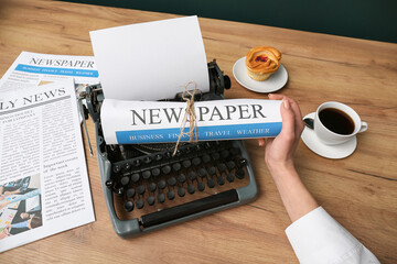 Female hand holding newspaper and cup of coffee with vintage typewriter on wooden table