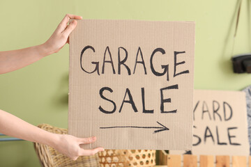 Woman holding cardboard with text GARAGE SALE in room of unwanted stuff