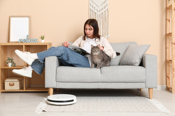 Young woman resting on sofa with cat and robot vacuum cleaner