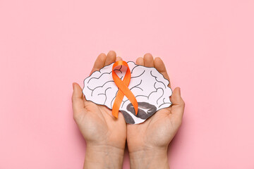 Female hands with orange ribbon and paper brain on pink background. Multiple Sclerosis Awareness...