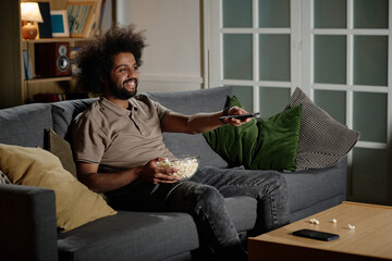 Young biracial guy sitting relaxed on couch in living room switching TV channels, copy space